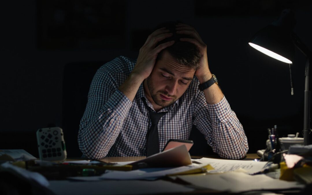 Man at desk with head in hands looking at phone.